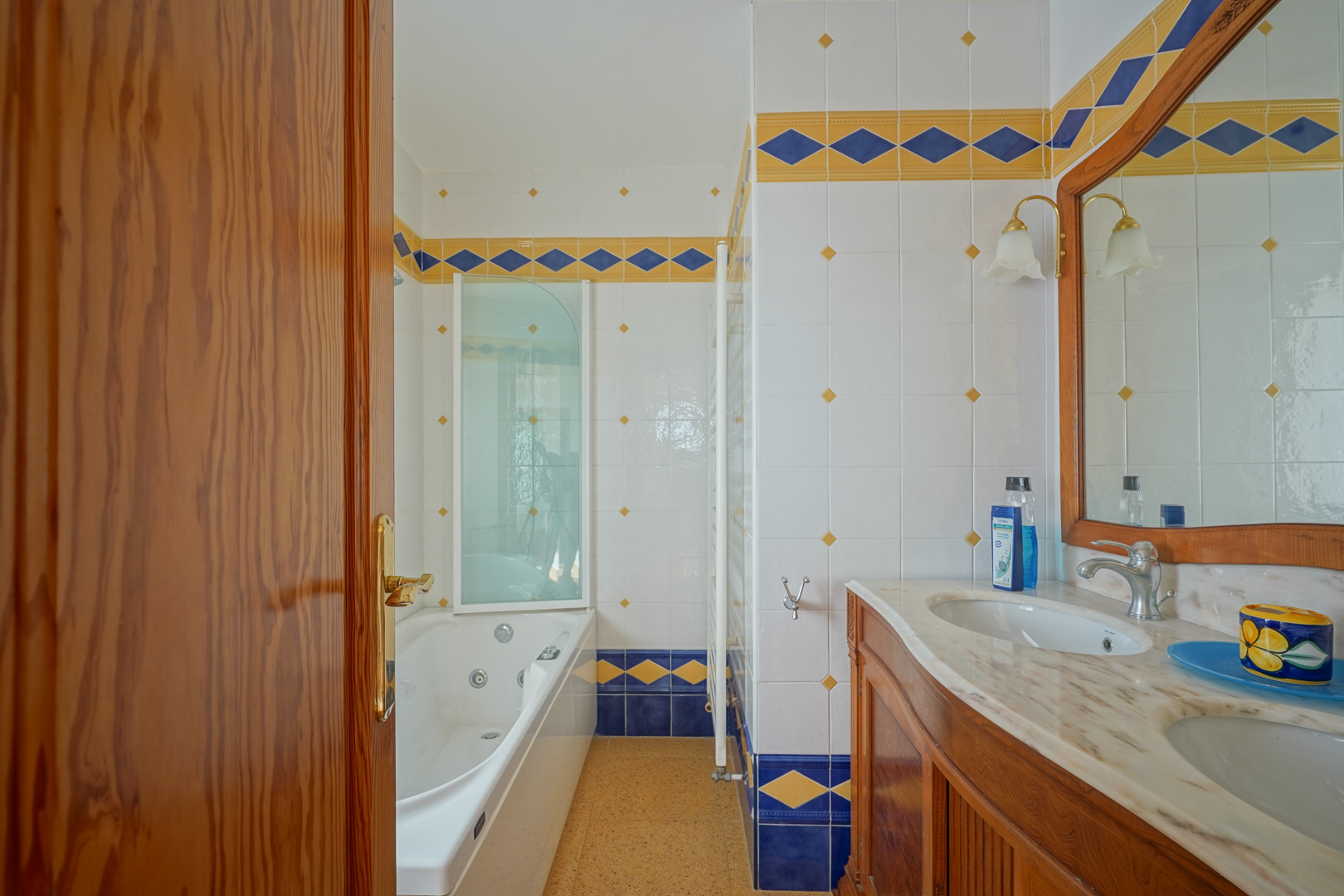 Cuarto de baño con lavabo doble de mármol sobre mueble de madera, bañera de hidromasaje y azulejos decorativos en tonos azul y amarillo de estilo mediterráneo.