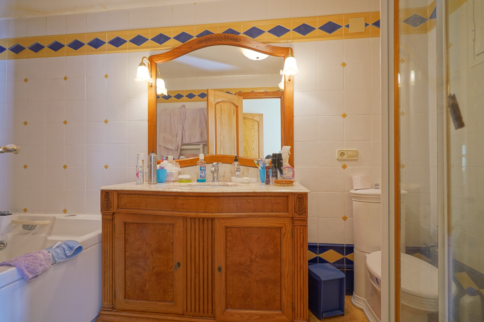 Cuarto de baño de estilo tradicional con mueble de madera tallada, encimera de mármol y azulejos decorativos con cenefas en azul y amarillo.