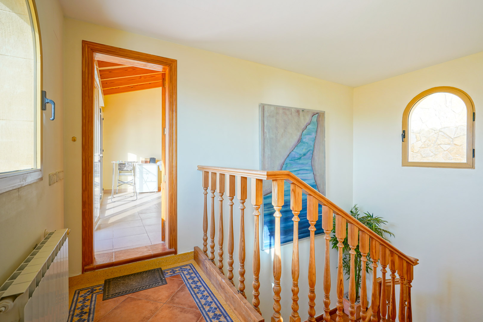 Distribuidor con escalera de madera, suelos cerámicos decorados y ventanal de arco. Conecta con una estancia luminosa de techos de madera y vigas vistas.