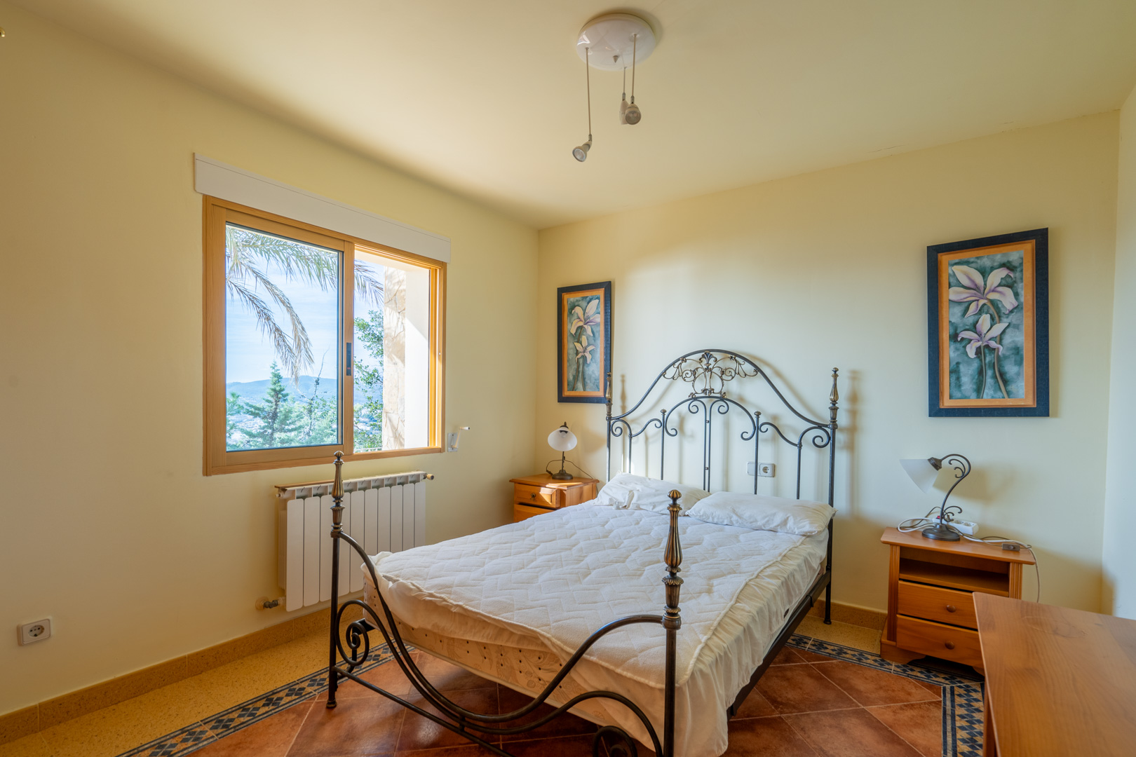 Dormitorio con cama de forja, suelos de terracota con cenefa decorativa y ventana de madera con vistas al paisaje mediterráneo de Jávea.
