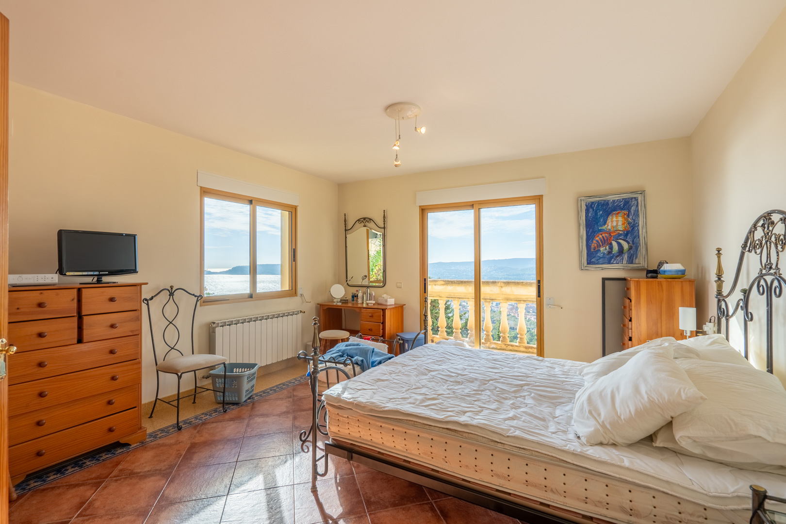 Dormitorio con suelos de terracota, cama de forja y acceso directo a un balcón con balaustrada que ofrece vistas panorámicas al mar y a la bahía de Jávea.