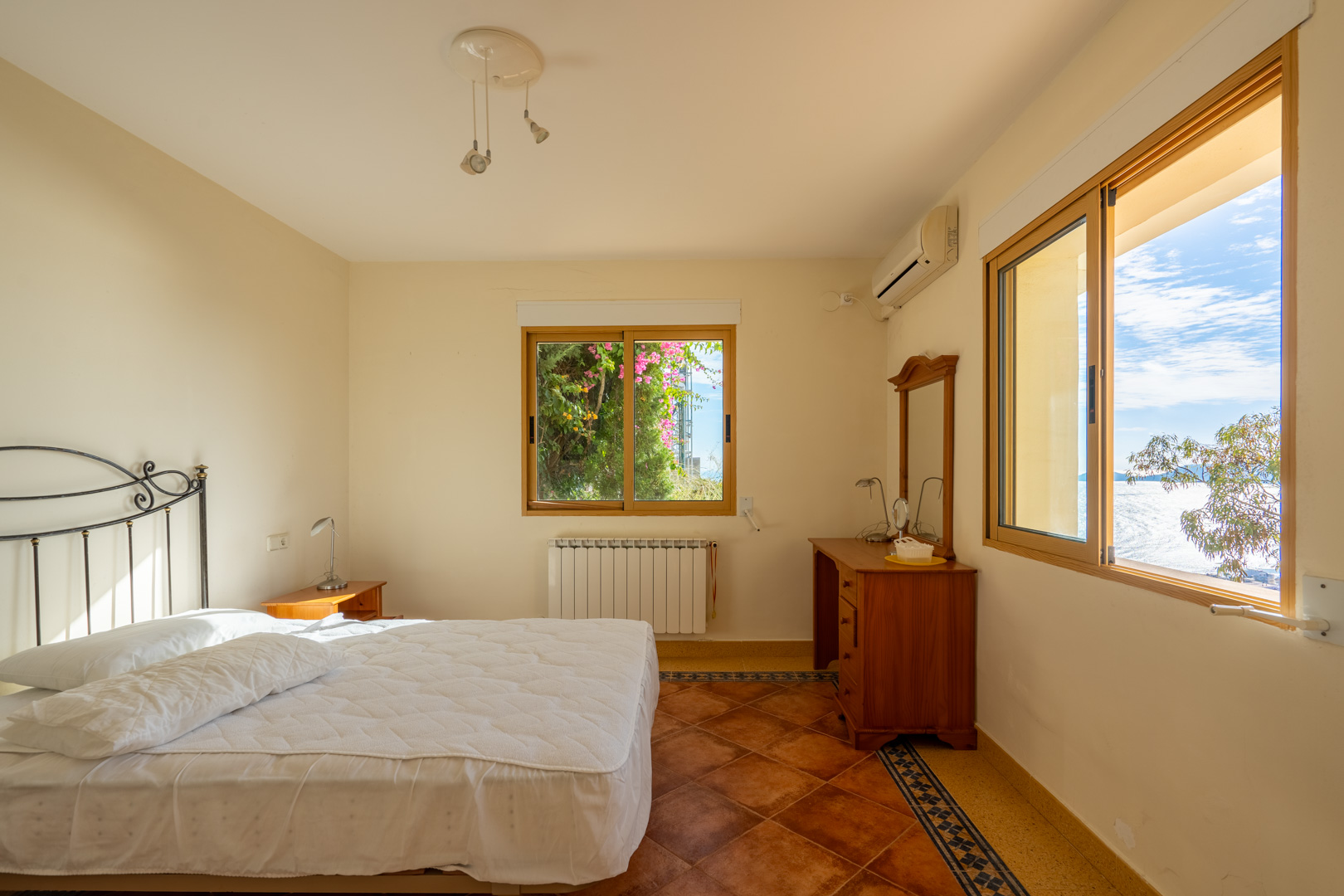 Dormitorio con suelos de terracota y cenefa decorativa. Incluye ventanas de madera con vistas al mar, cabecero de forja y aire acondicionado split.