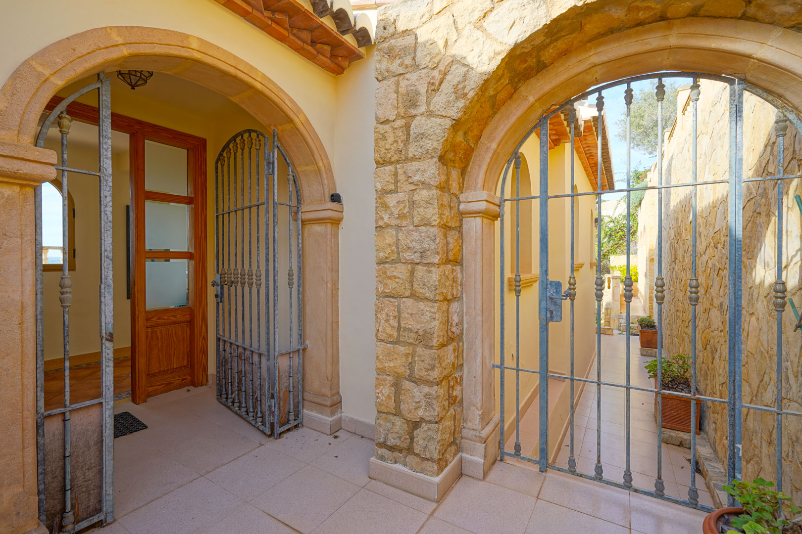 Entrada con arcos de piedra tosca y rejas de hierro forjado que conducen a la puerta principal de madera y a un pasillo lateral pavimentado.