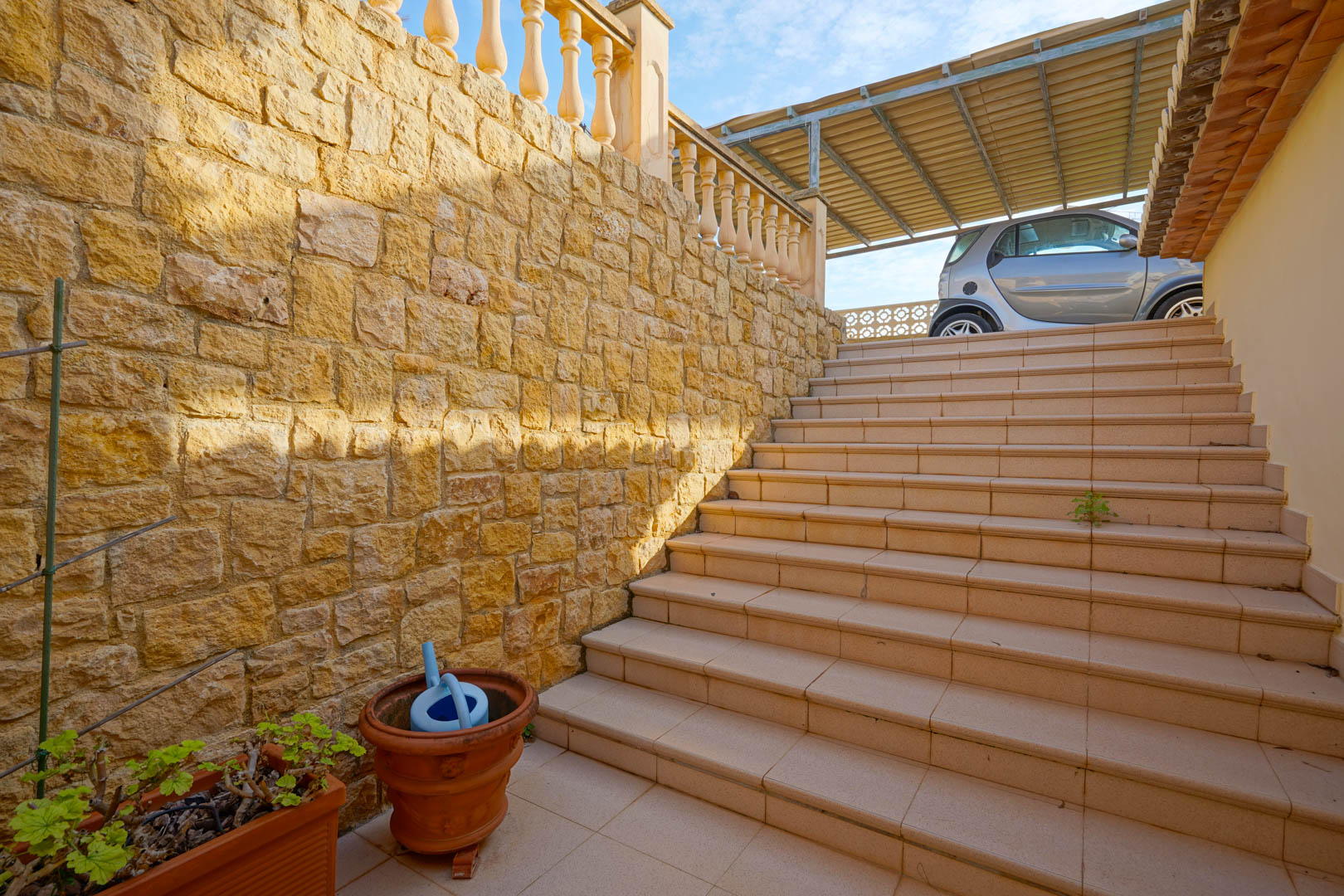 Escalera exterior de gres flanqueada por un muro de piedra natural, que conecta los niveles de la villa con la zona de aparcamiento cubierta.