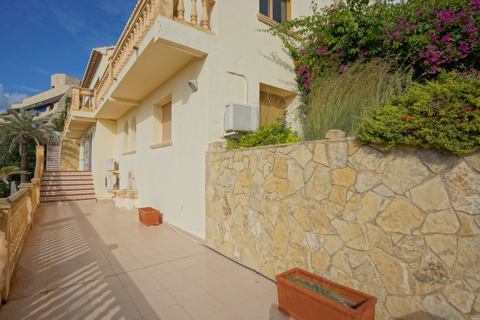 Fachada lateral con muro de piedra natural, suelos cerámicos y escaleras decoradas con azulejos tradicionales. Incluye jardineras de terracota y vegetación mediterránea.