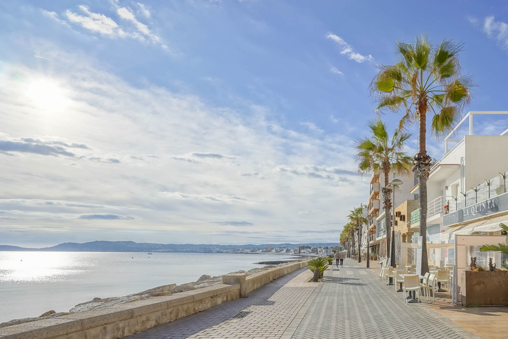 Paseo marítimo de Jávea con palmeras y terrazas frente al mar, ofreciendo vistas despejadas de la bahía y el perfil montañoso bajo un cielo soleado.