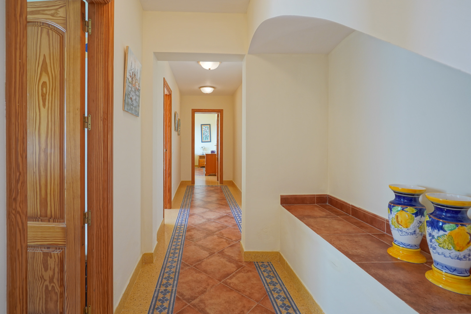 Pasillo con suelos de baldosas de terracota y cenefas azules, puertas de madera maciza y un banco de obra decorado con jarrones cerámicos tradicionales.