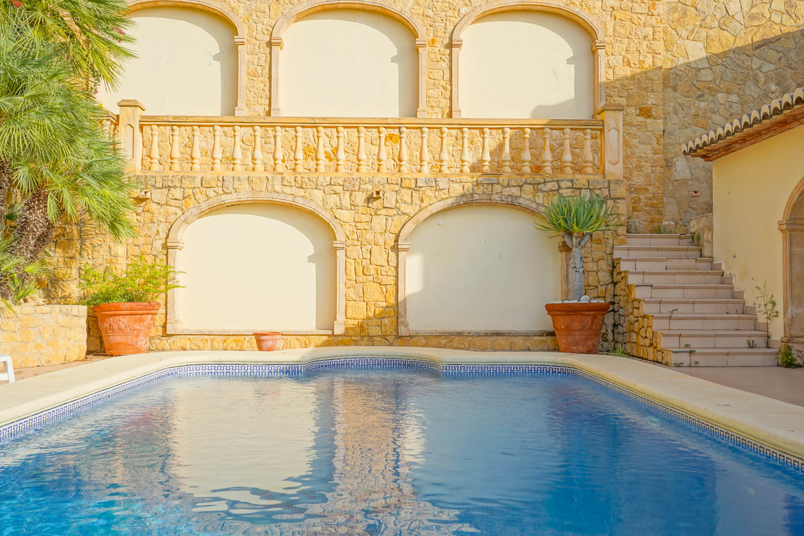 Piscina con borde de gresite azul y cenefa clásica, flanqueada por muros de piedra natural con nichos arqueados, balustrada superior y vegetación mediterránea en macetas.