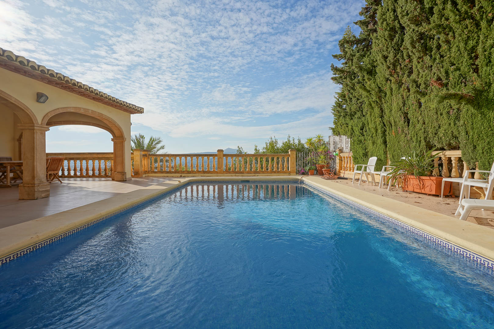 Piscina rectangular con borde de piedra, situada junto a un porche de arcos tradicionales y una balaustrada clásica con vistas al mar y cipreses laterales.