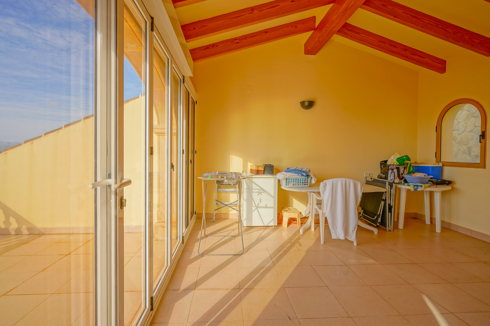 Terraza acristalada con vigas de madera a la vista, suelos cerámicos y amplios ventanales correderos que permiten la entrada de abundante luz natural.