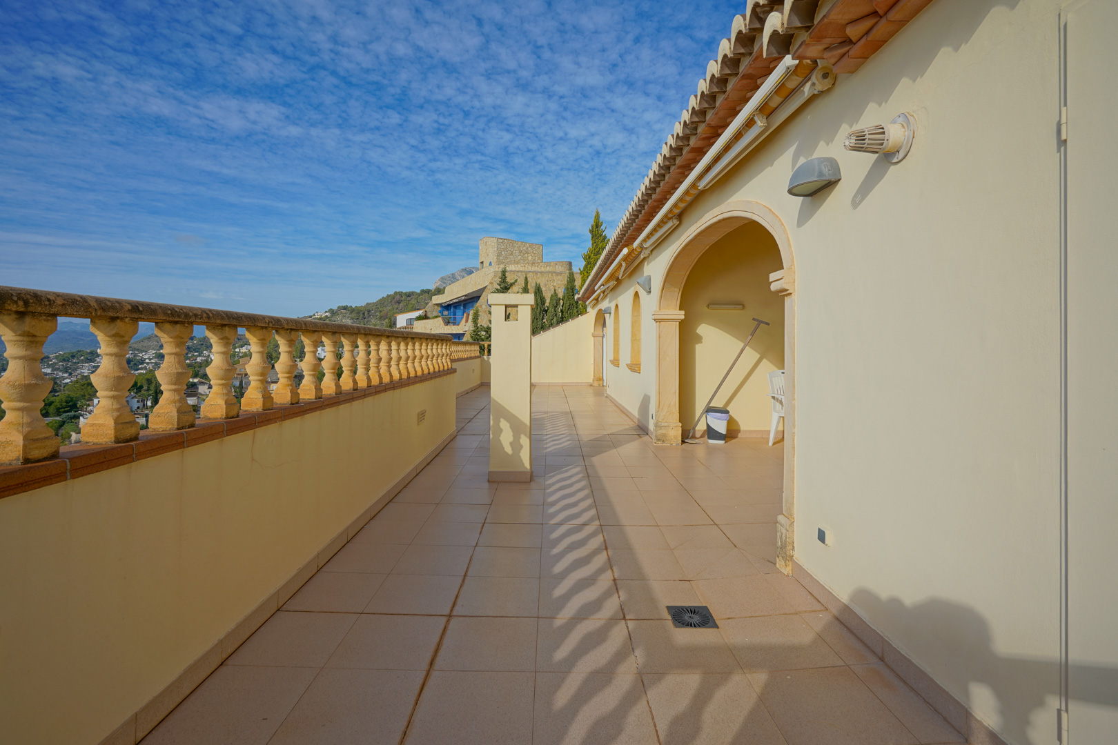 Terraza alargada con suelo cerámico y balaustrada tradicional que ofrece vistas despejadas al valle. La fachada presenta arcos de medio punto y tejado de teja clásica.