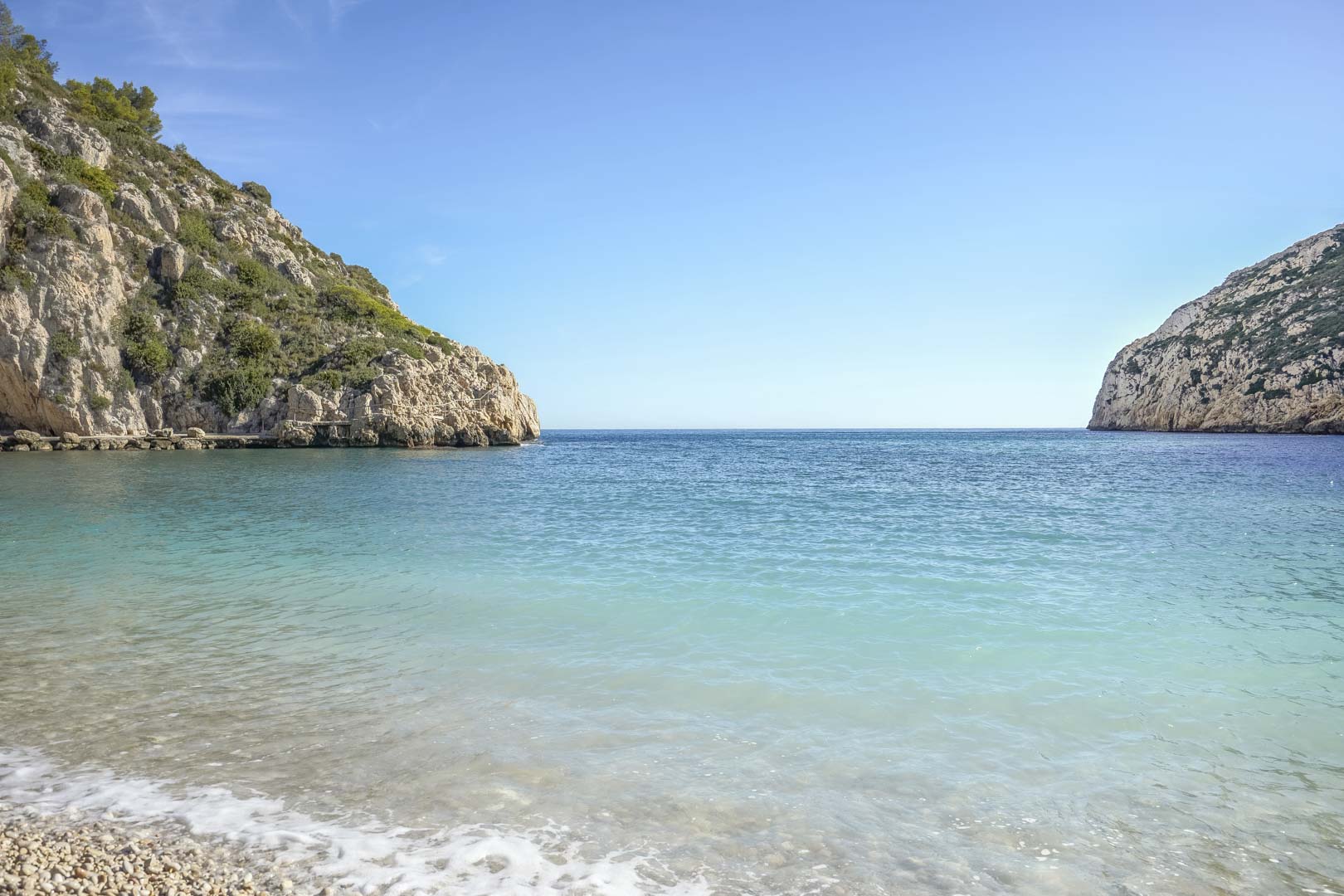 Vista de una cala de aguas cristalinas y turquesas con orilla de guijarros, flanqueada por acantilados mediterráneos bajo un cielo despejado en Jávea.