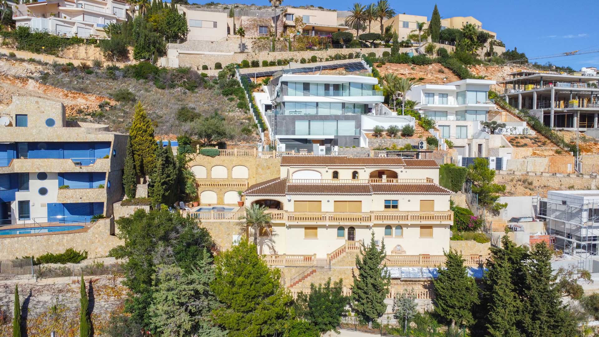 Vista exterior de la villa tradicional en La Corona, destacando su fachada color crema, tejados de teja cerámica y niveles de terrazas con balaustradas integradas en la ladera.