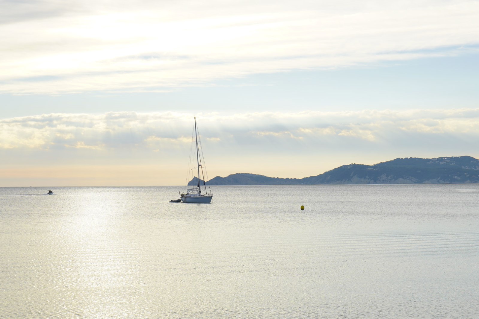 Vista panorámica de la bahía de Jávea con un velero en aguas tranquilas, destacando el perfil del Cap Negre bajo la suave luz del amanecer.