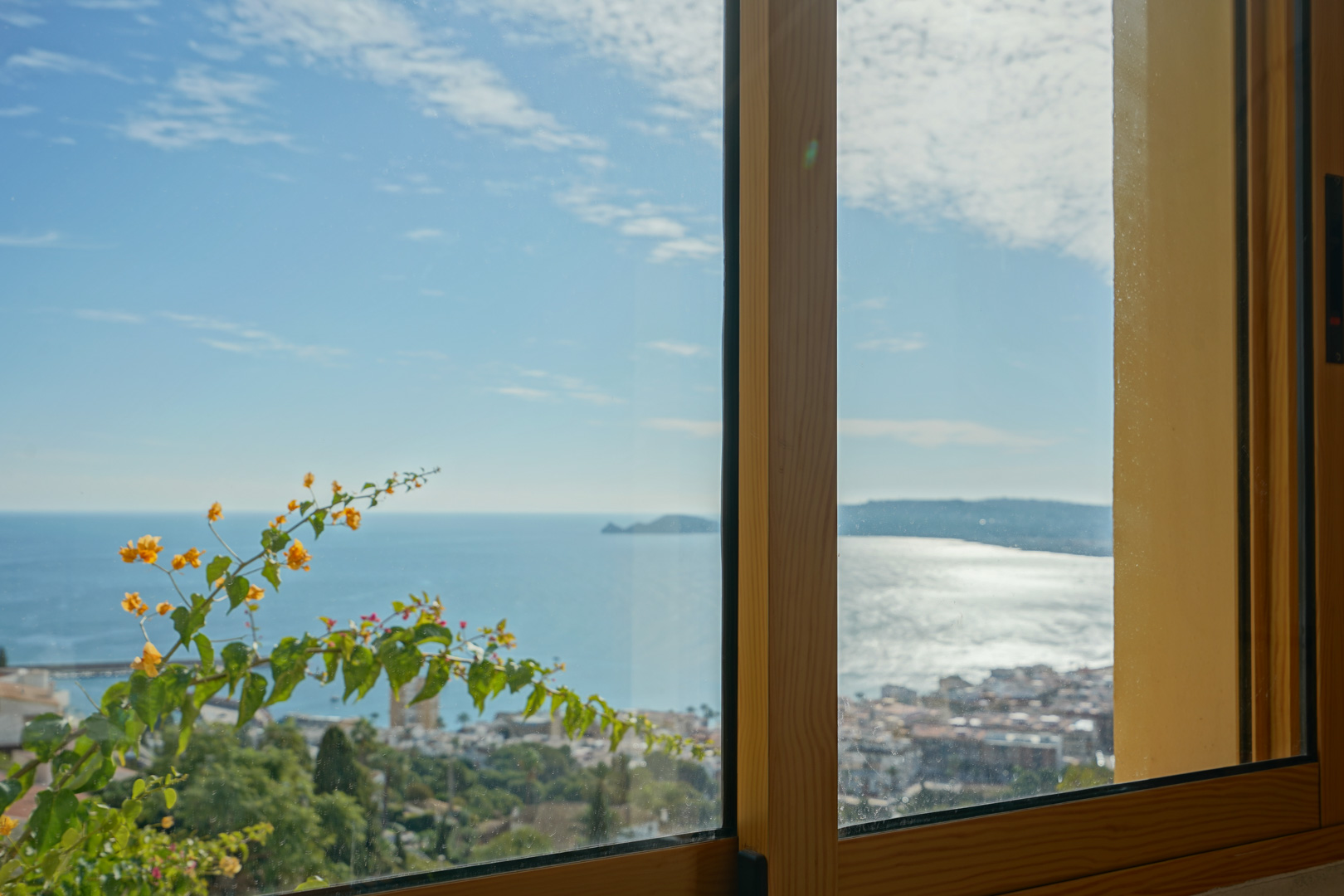 Vista panorámica de la bahía de Jávea y el Cap Negre a través de un ventanal con marco de madera, decorada por buganvillas en flor.