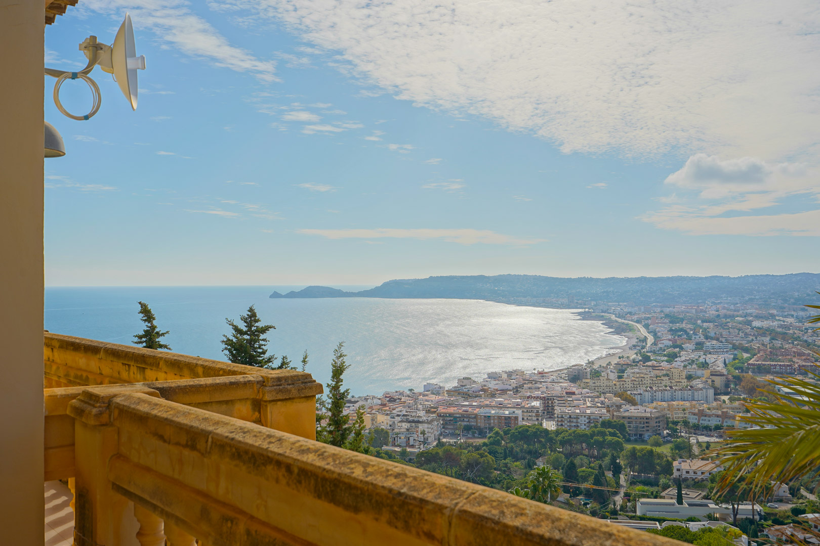 Vista panorámica desde la terraza con balaustrada de piedra, capturando la bahía de Jávea, el puerto y el perfil del Cap Negre bajo un cielo despejado.