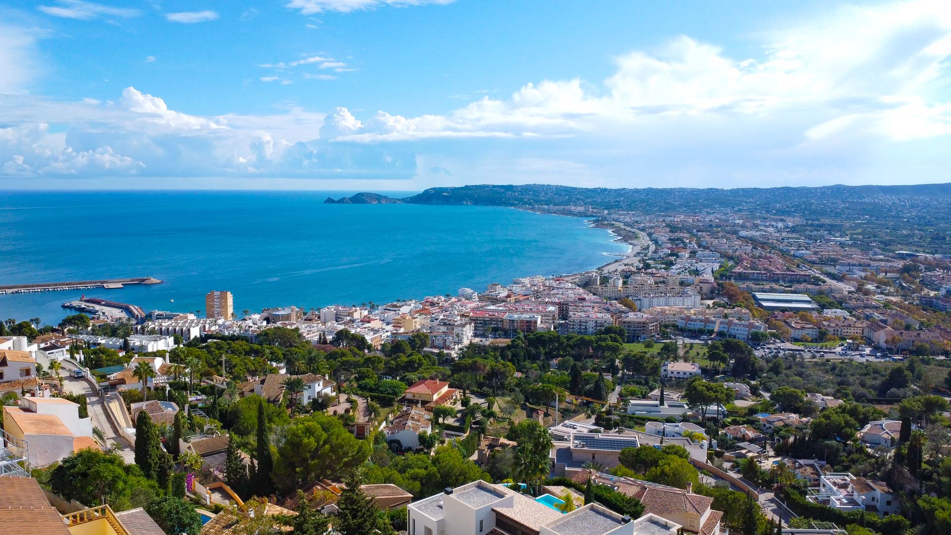 Vista panorámica desde la urbanización La Corona que abarca la bahía de Jávea, el puerto pesquero y el cabo Negre bajo un cielo despejado.