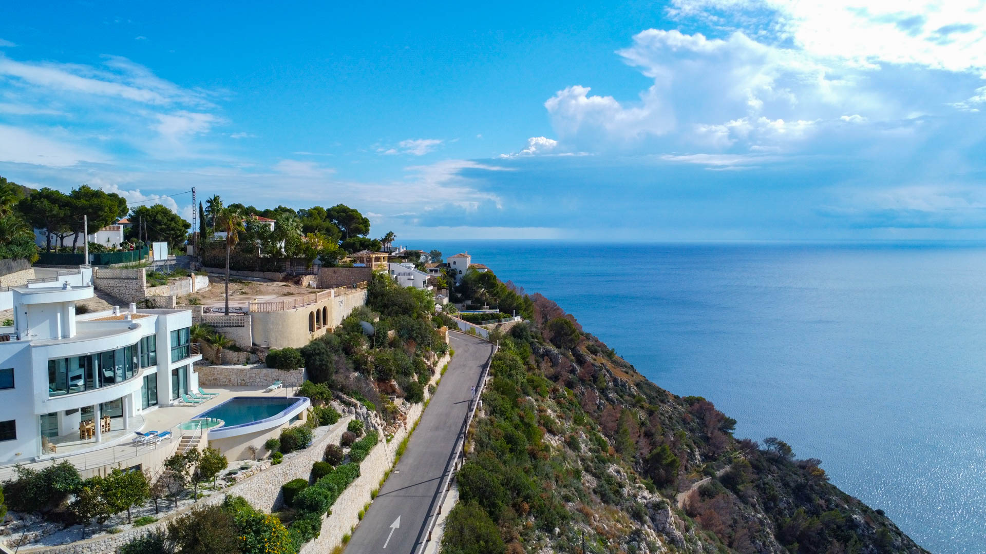 Vista aérea de la parcela elevada con vistas panorámicas al mar Mediterráneo, situada junto a villas de diseño moderno y una carretera costera de acceso.