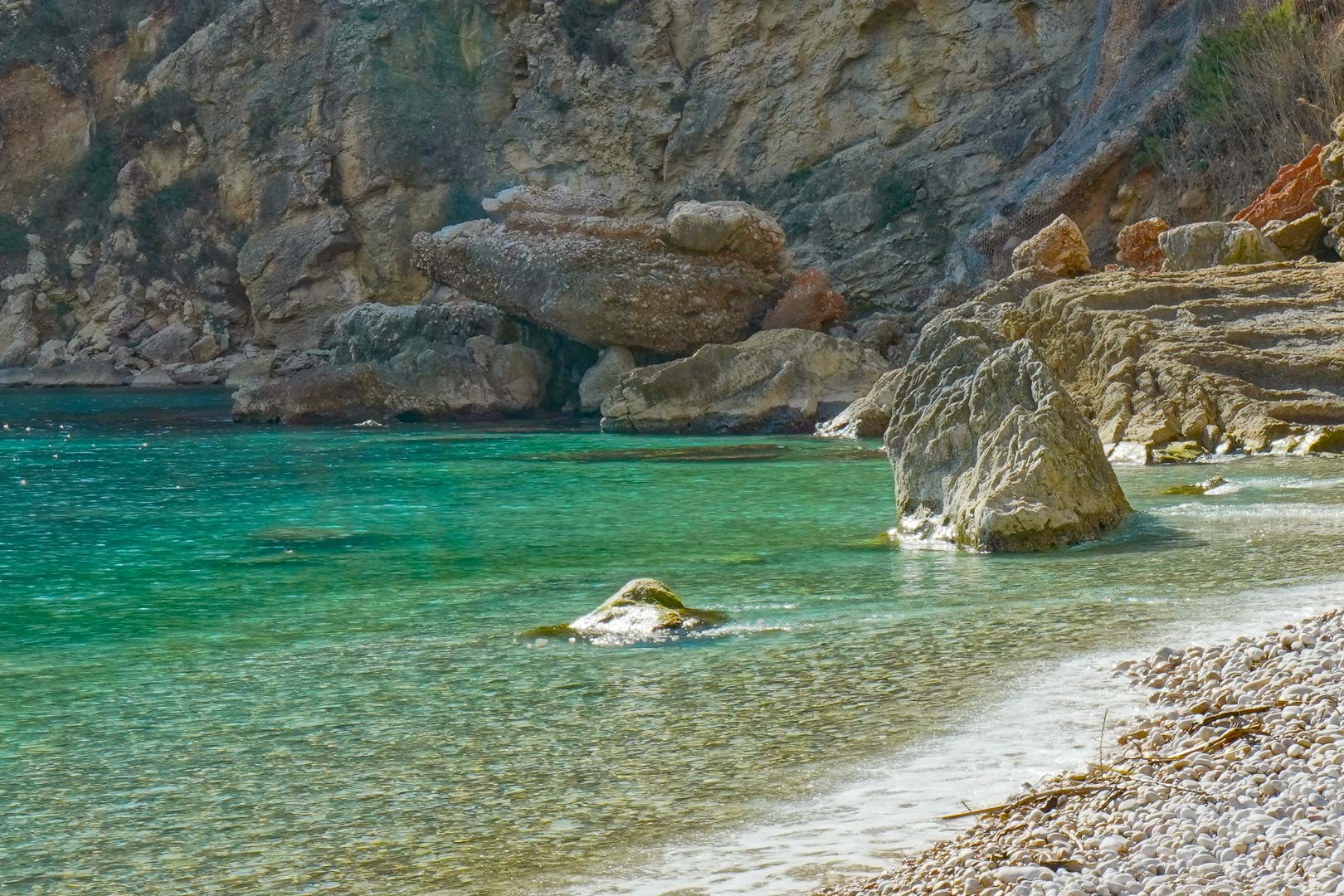 Vista de la orilla de guijarros en la Cala Granadella, destacando sus aguas cristalinas de tono turquesa y las formaciones rocosas naturales del entorno.