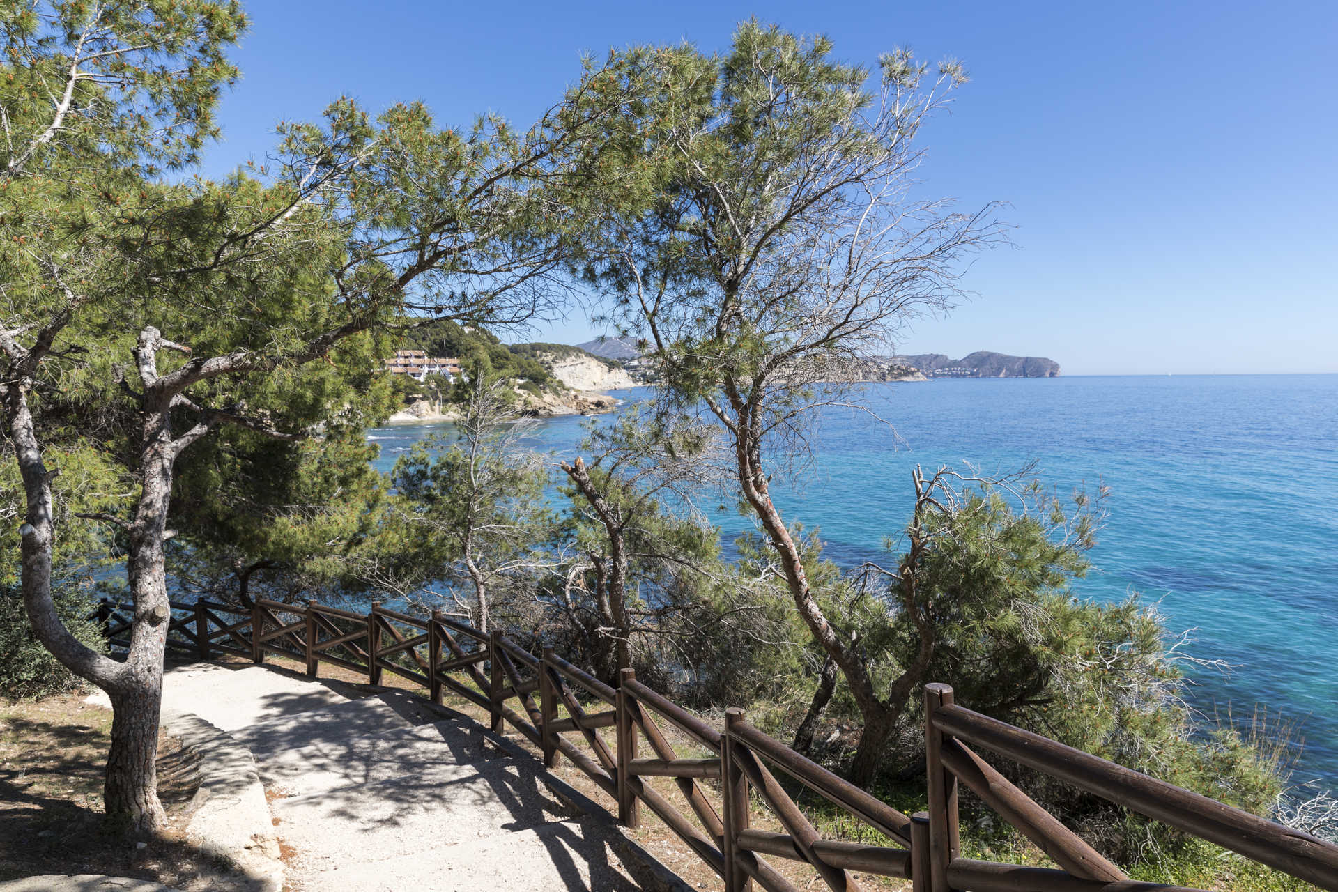 Sendero costero con barandilla de madera y pinos mediterráneos, ofreciendo vistas panorámicas a las aguas turquesas cerca de la playa de La Fustera.