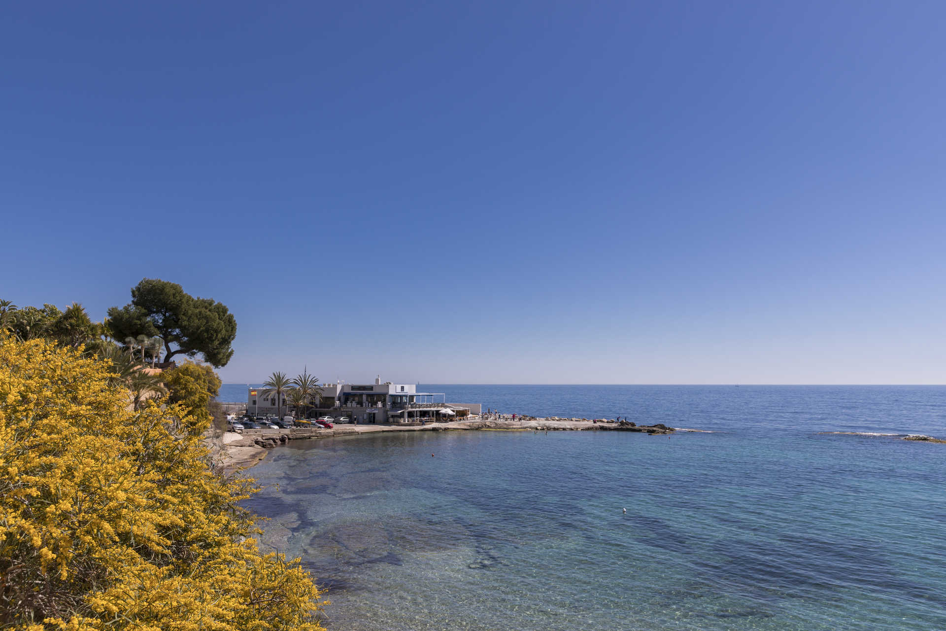 Vista panorámica de la cala de La Fustera con aguas cristalinas, vegetación mediterránea en primer plano y un restaurante costero bajo el cielo despejado.