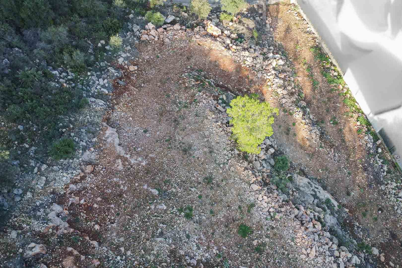 Vista aérea de la parcela nivelada con terreno rocoso y vegetación autóctona. Se observa el vallado perimetral y la preparación del suelo para la construcción.