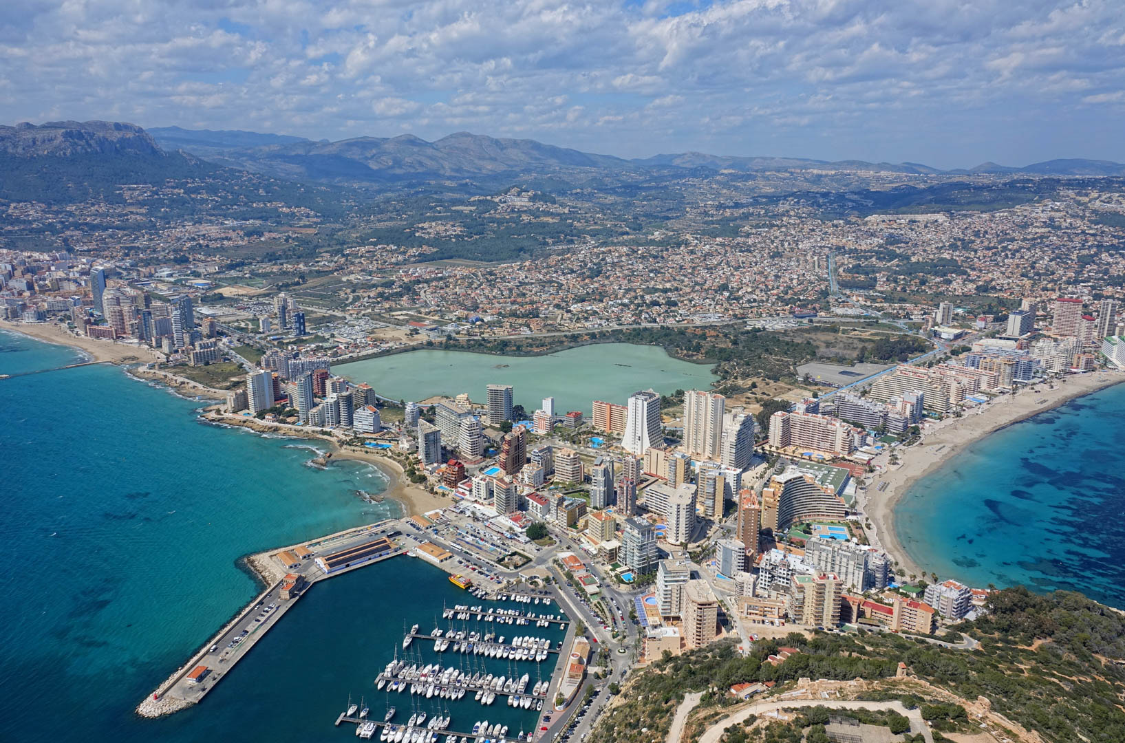Vista aérea panorámica de Calpe que muestra el puerto deportivo, las playas de arena, la laguna de Las Salinas y el perfil urbano con las montañas al fondo.