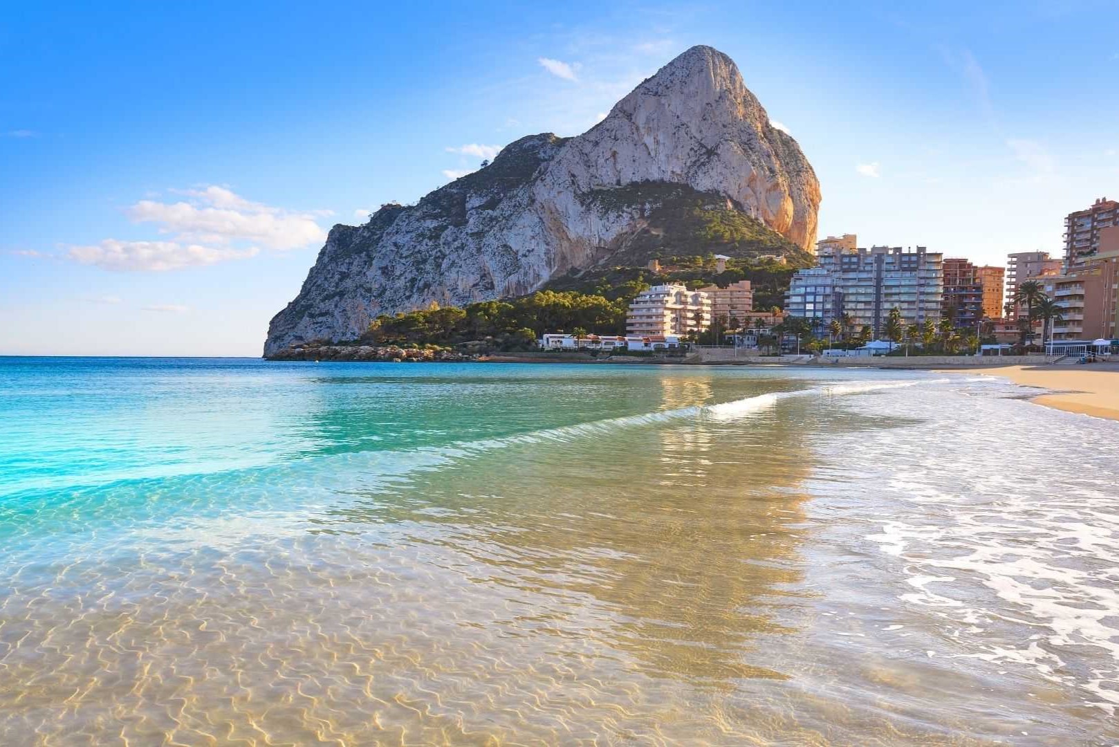 Vista panorámica del Peñón de Ifach desde la orilla de la playa en Calpe, destacando las aguas cristalinas del Mediterráneo y el perfil costero bajo un cielo despejado.