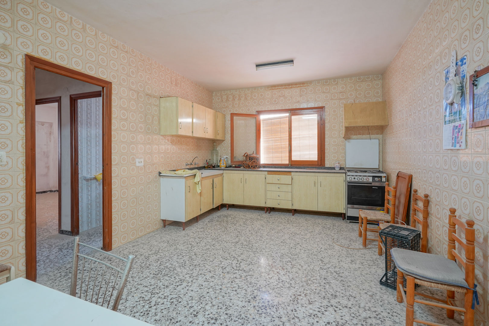 Cocina espaciosa con azulejos decorativos de época, suelos de terrazo y mobiliario de madera clara, iluminada por luz natural a través de su ventana central.