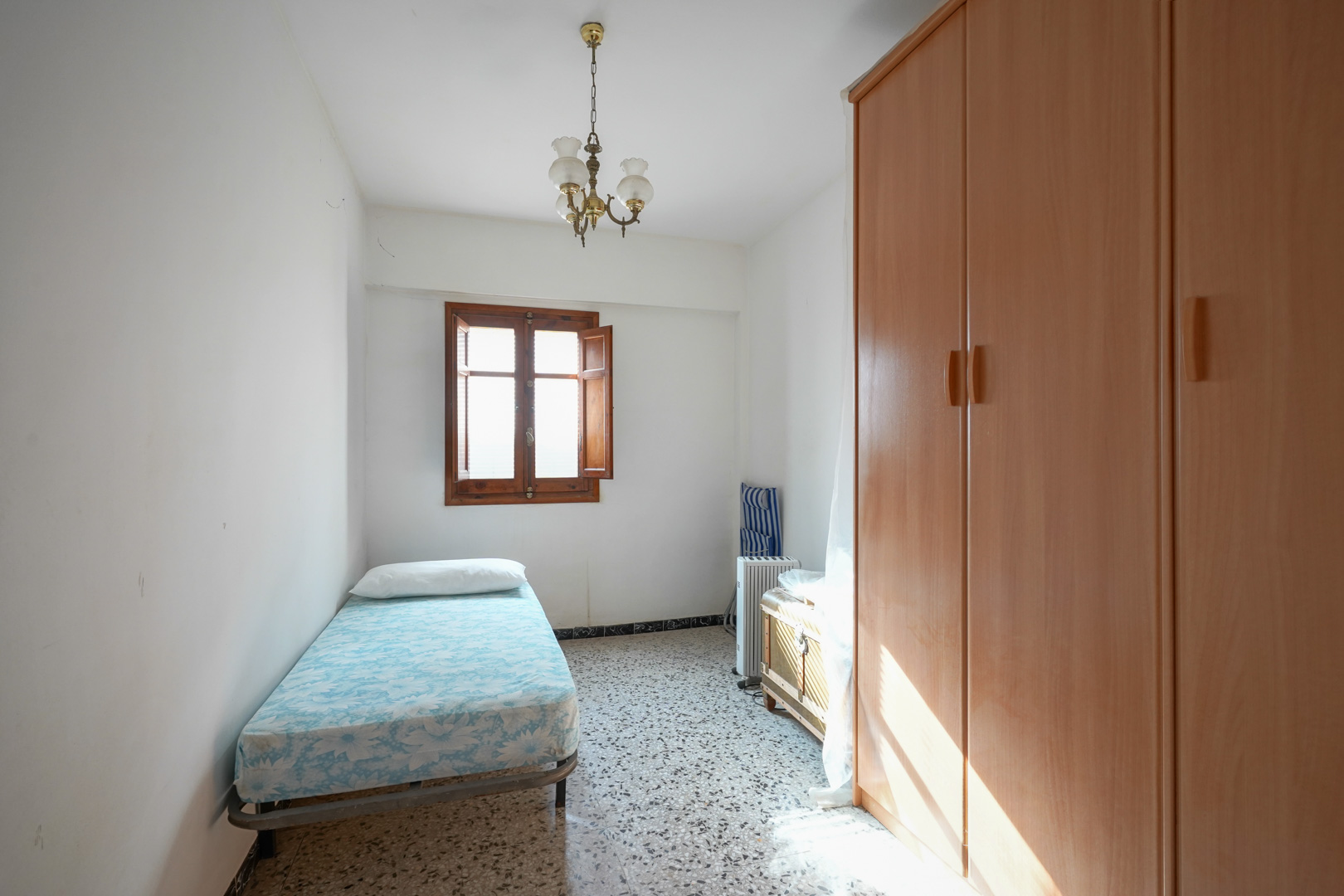 Dormitorio individual con suelos de terrazo, armario de madera de gran tamaño y ventana con contraventanas de madera que aporta luz natural.