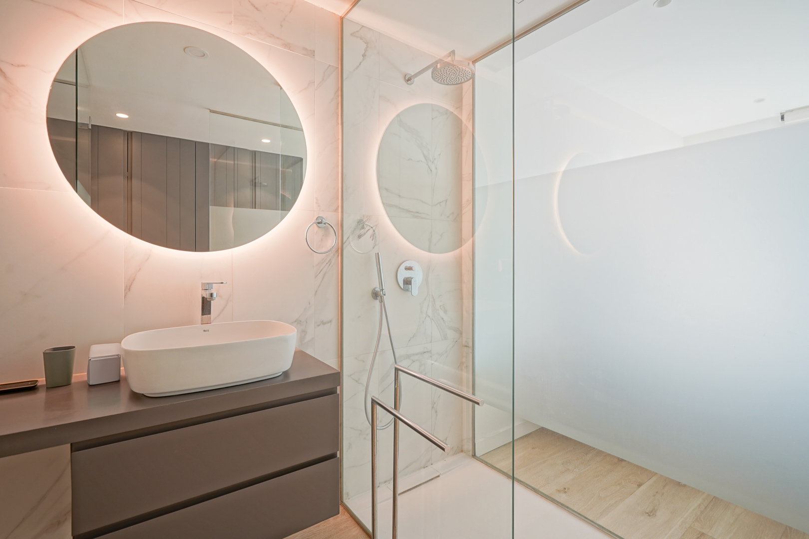 Baño moderno con lavabo sobre encimera, espejo circular retroiluminado y ducha de obra con mampara de cristal y revestimiento cerámico efecto mármol.