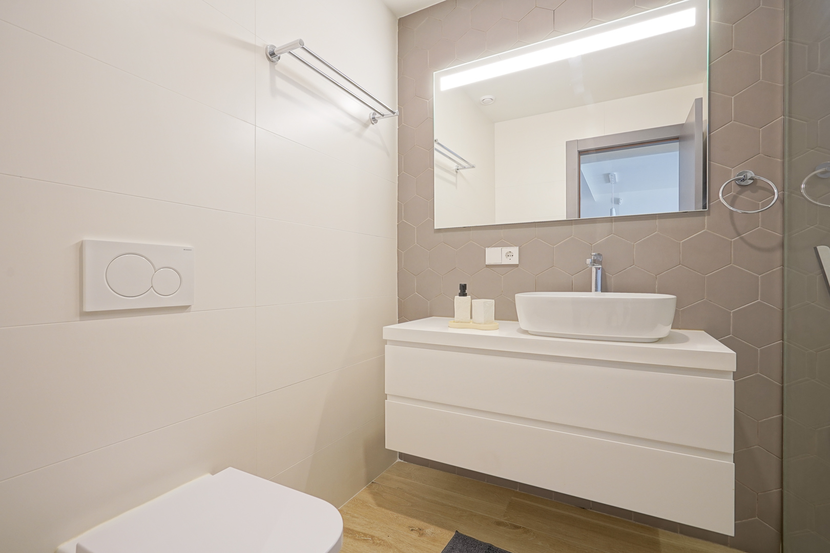 Cuarto de baño moderno con azulejos hexagonales en tono topo, lavabo sobre encimera y mueble suspendido blanco. Incluye espejo con iluminación LED integrada y grifería cromada.