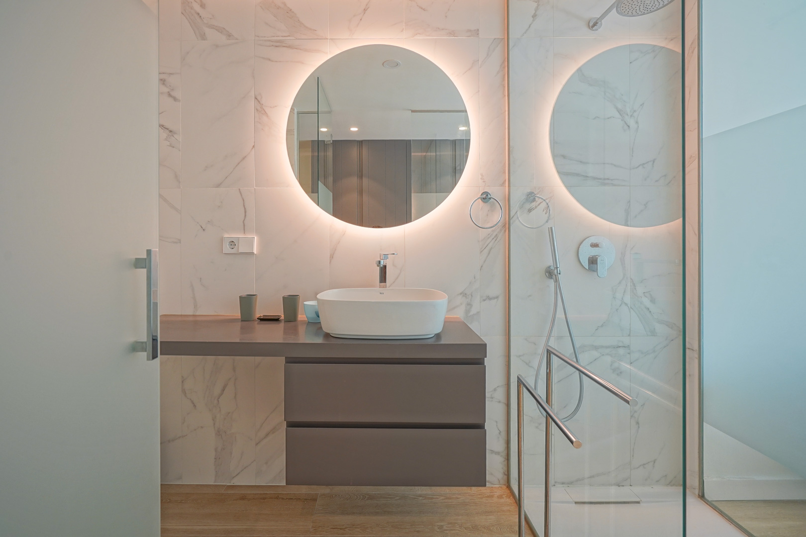 Cuarto de baño moderno con lavabo sobre encimera, espejo circular retroiluminado y paredes revestidas en gres cerámico efecto mármol.
