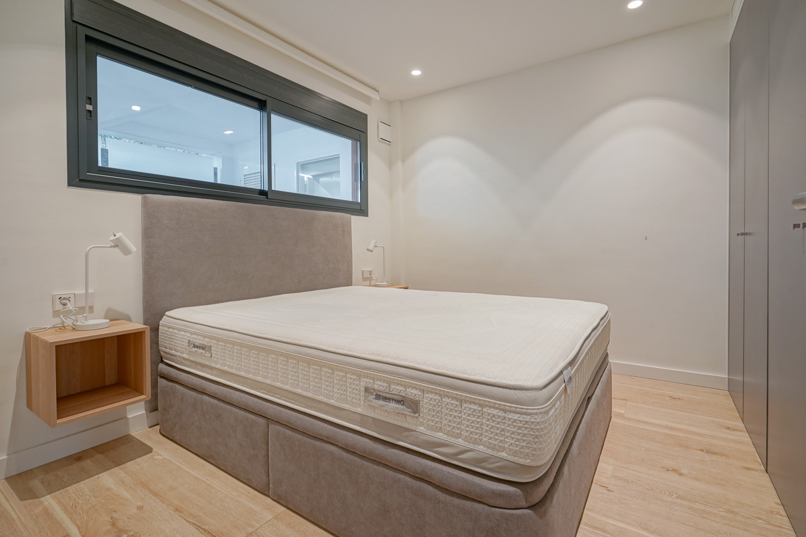 Dormitorio moderno con cama doble, cabecero tapizado, armarios empotrados y suelos de madera clara bajo iluminación empotrada.