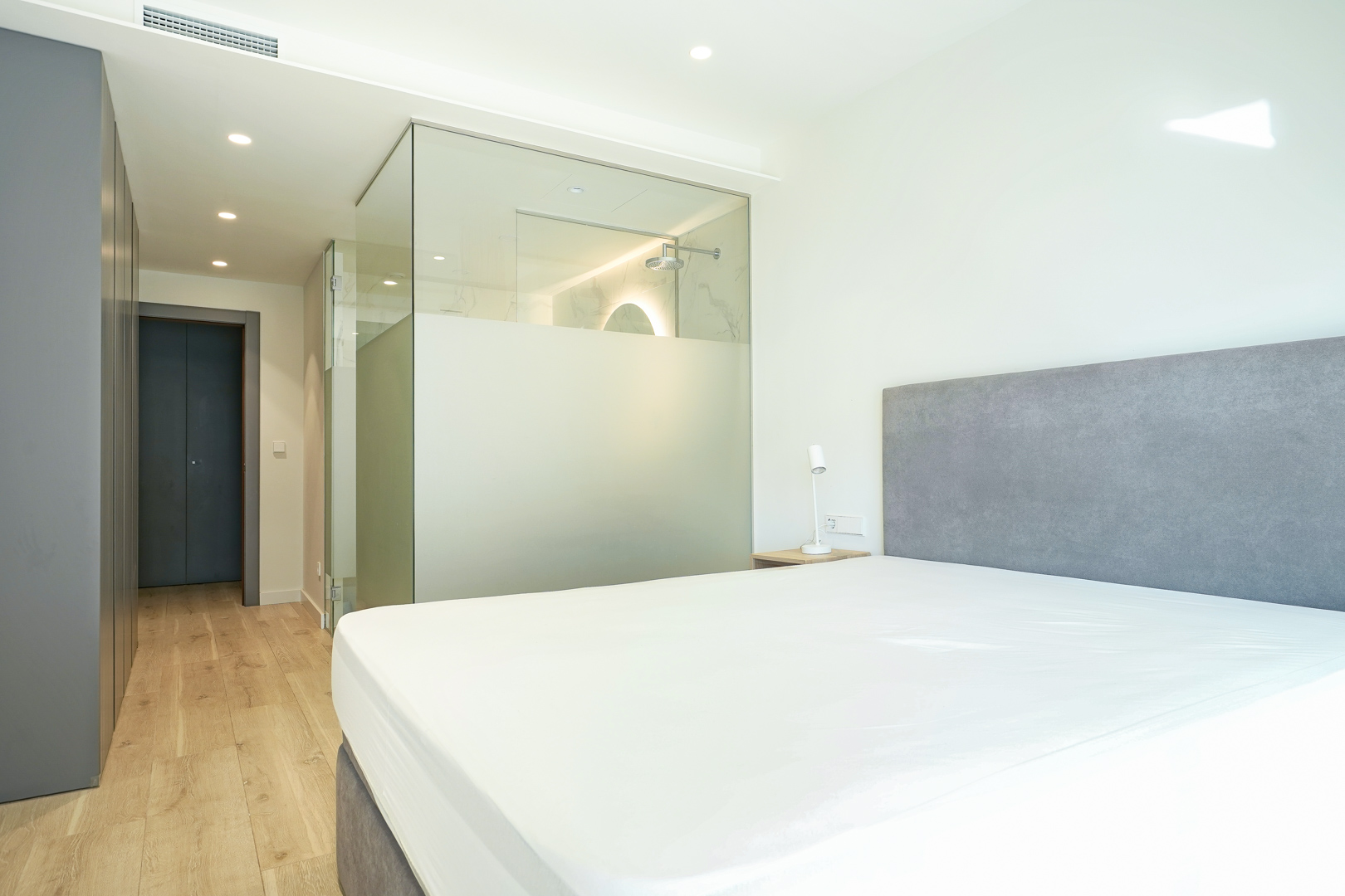 Dormitorio principal con baño en suite integrado mediante mampara de cristal translúcido, armarios empotrados y suelos de madera clara.