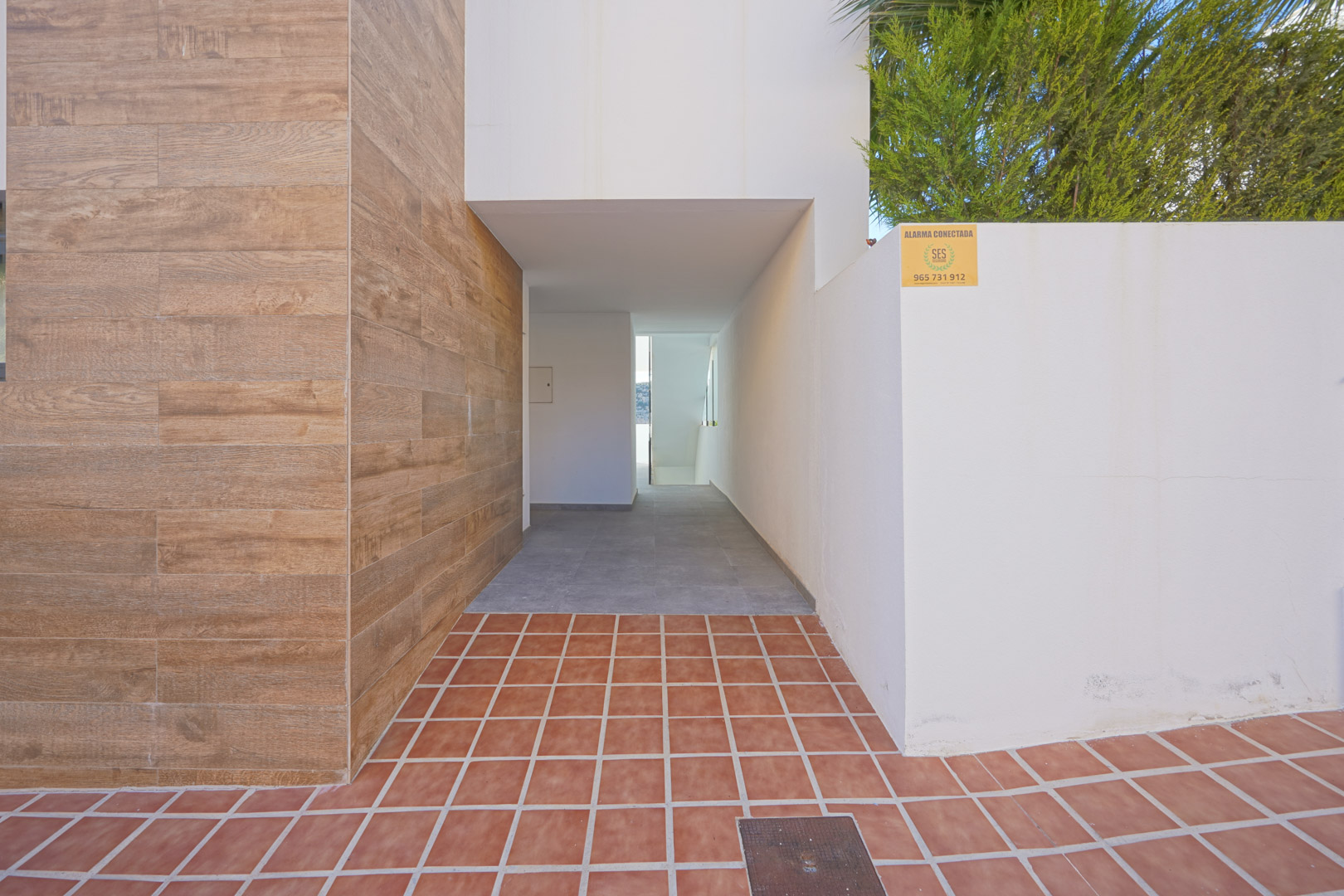 Entrada exterior con revestimiento cerámico de imitación madera, suelo de terracota y acceso cubierto hacia el interior del edificio Monte Cala Gardens.
