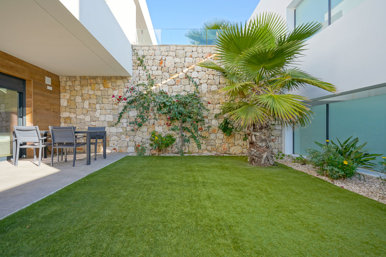 Jardín privado con césped artificial, una palmera y muro de piedra natural. Incluye una zona de comedor exterior bajo el porche cubierto con acabados modernos.
