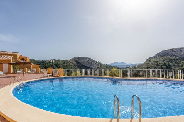 Piscina comunitaria de forma curva en Monte Cala Gardens, rodeada de una amplia terraza con tumbonas y vistas despejadas a las montañas del parque natural.