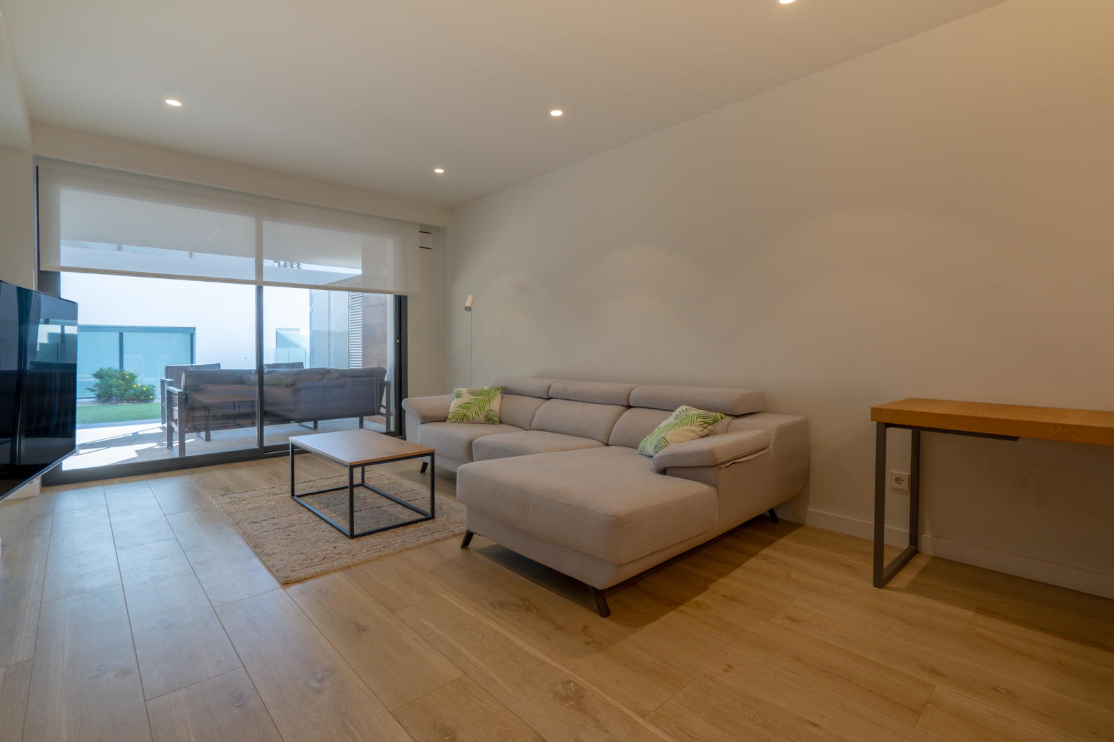 Salón moderno con sofá tipo chaise longue, suelos de madera clara y acceso directo a la terraza mediante amplios ventanales correderos con estores eléctricos.