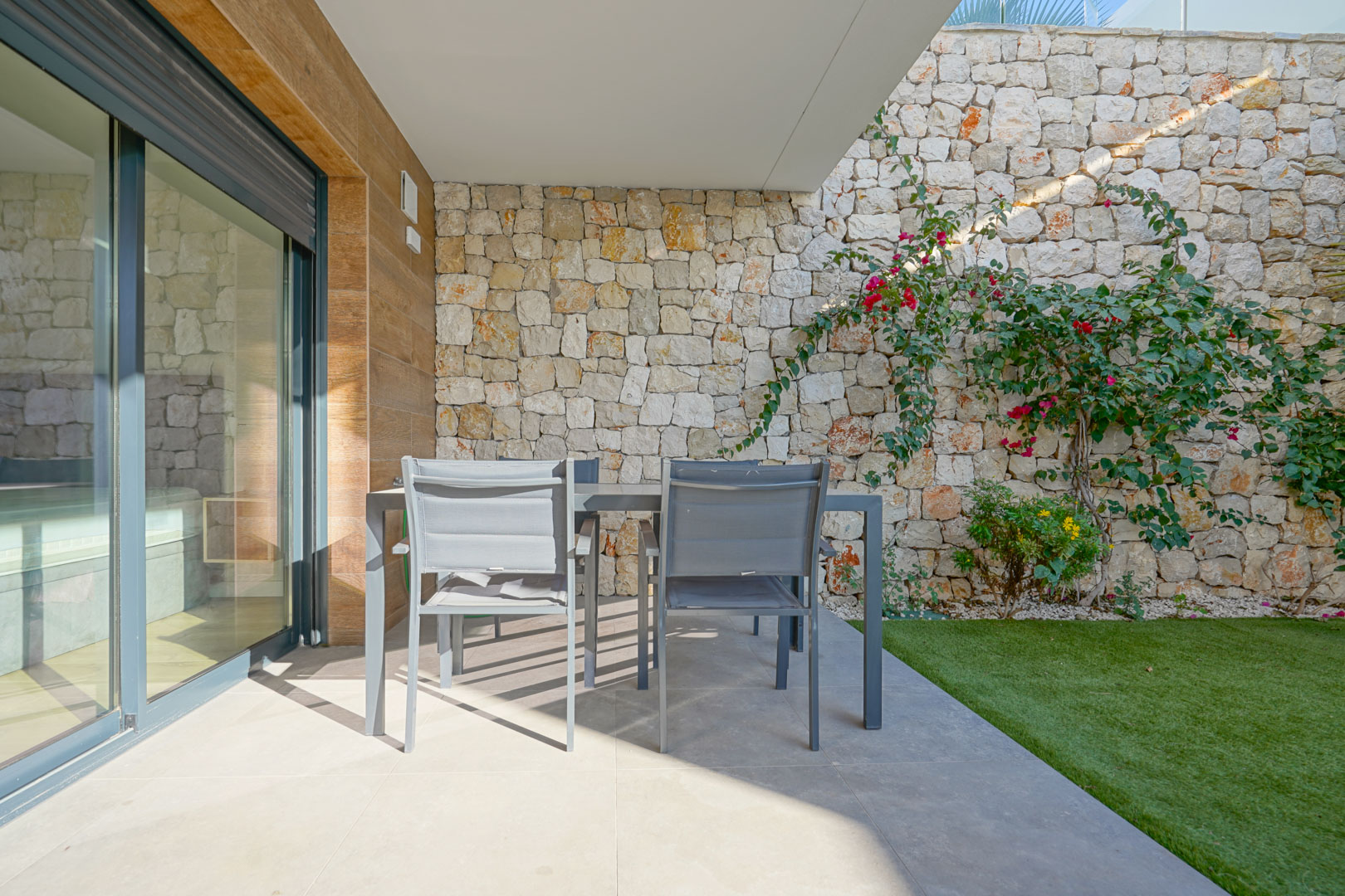 Terraza cubierta con comedor exterior, delimitada por muros de piedra natural y un jardín privado con césped artificial.