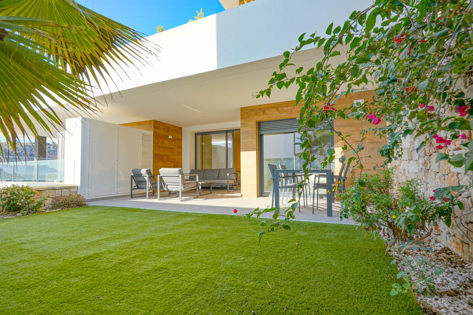 Terraza cubierta con mobiliario de exterior y jardín privado de césped artificial, delimitado por un muro de piedra natural y vegetación mediterránea.