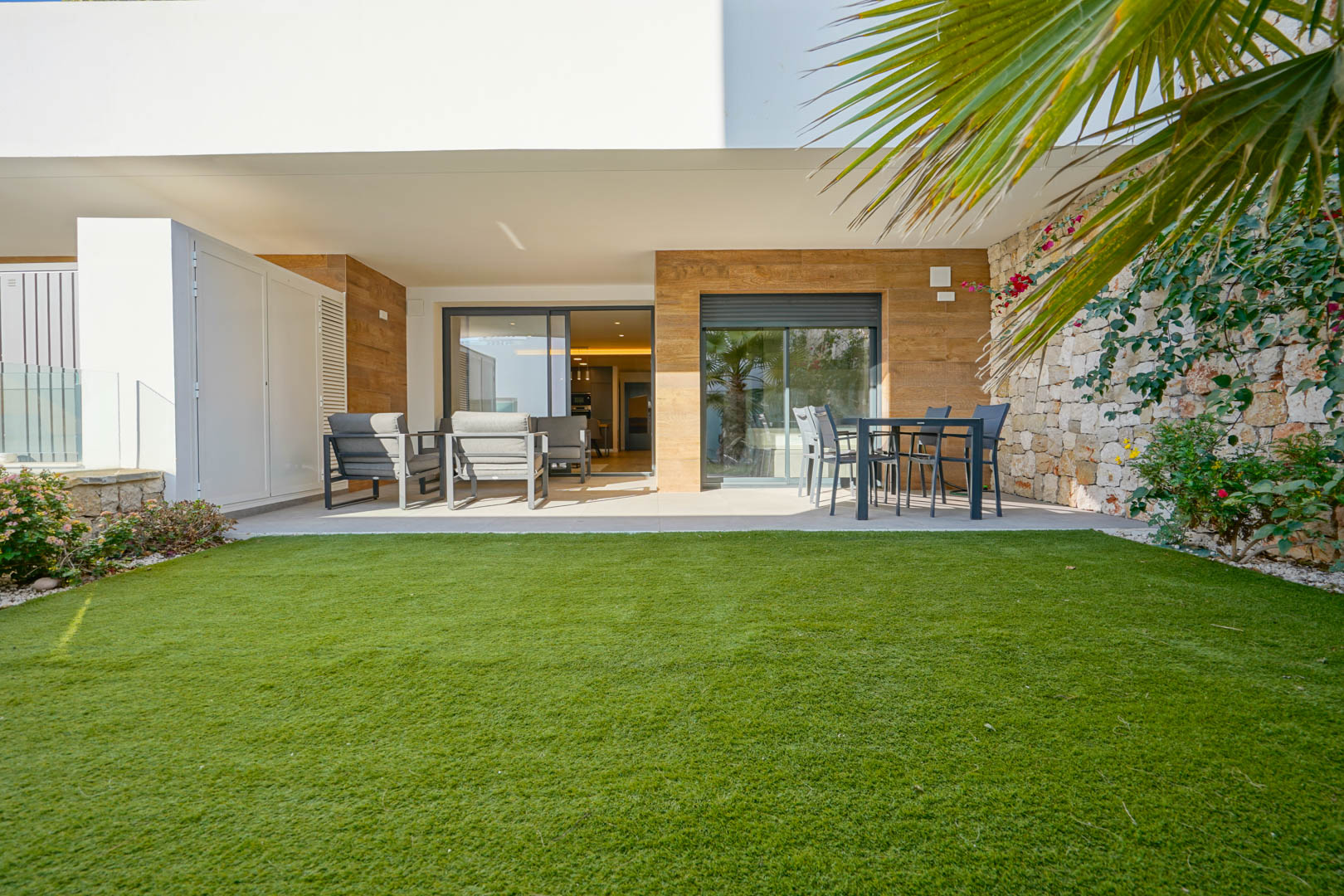 Terraza cubierta con mobiliario de exterior y jardín privado de césped, delimitado por un muro de piedra natural en una vivienda de estilo moderno.