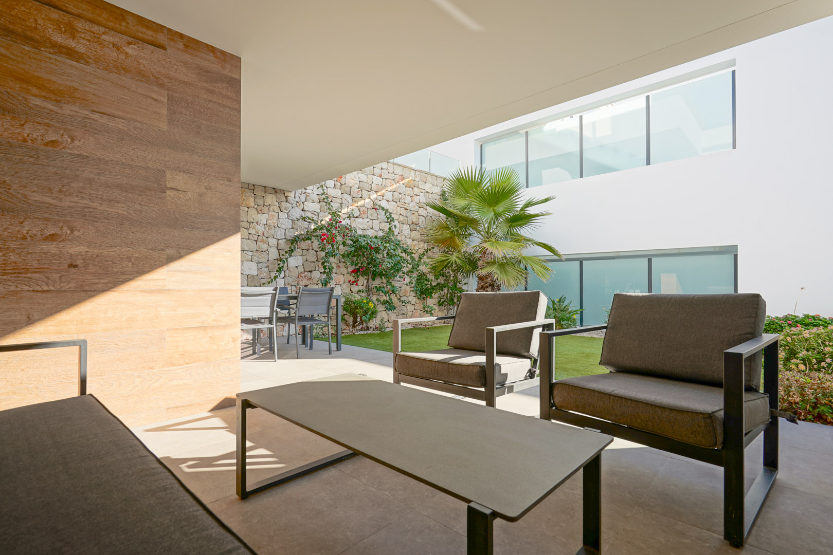 Terraza cubierta con zona de estar y comedor, pared de piedra natural y jardín privado con palmeras en Monte Cala Gardens.