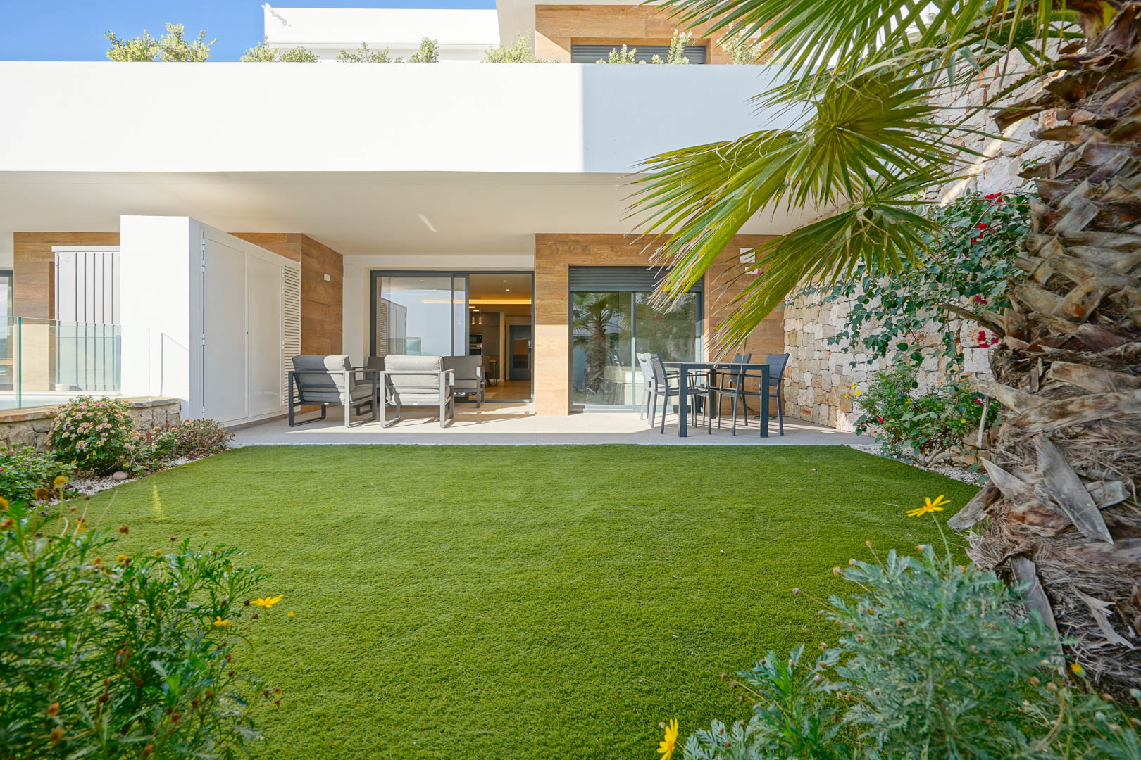 Terraza cubierta de 23 m2 y jardín privado de 36 m2 con césped artificial, delimitado por un muro de piedra natural y acceso directo al salón y dormitorio principal.