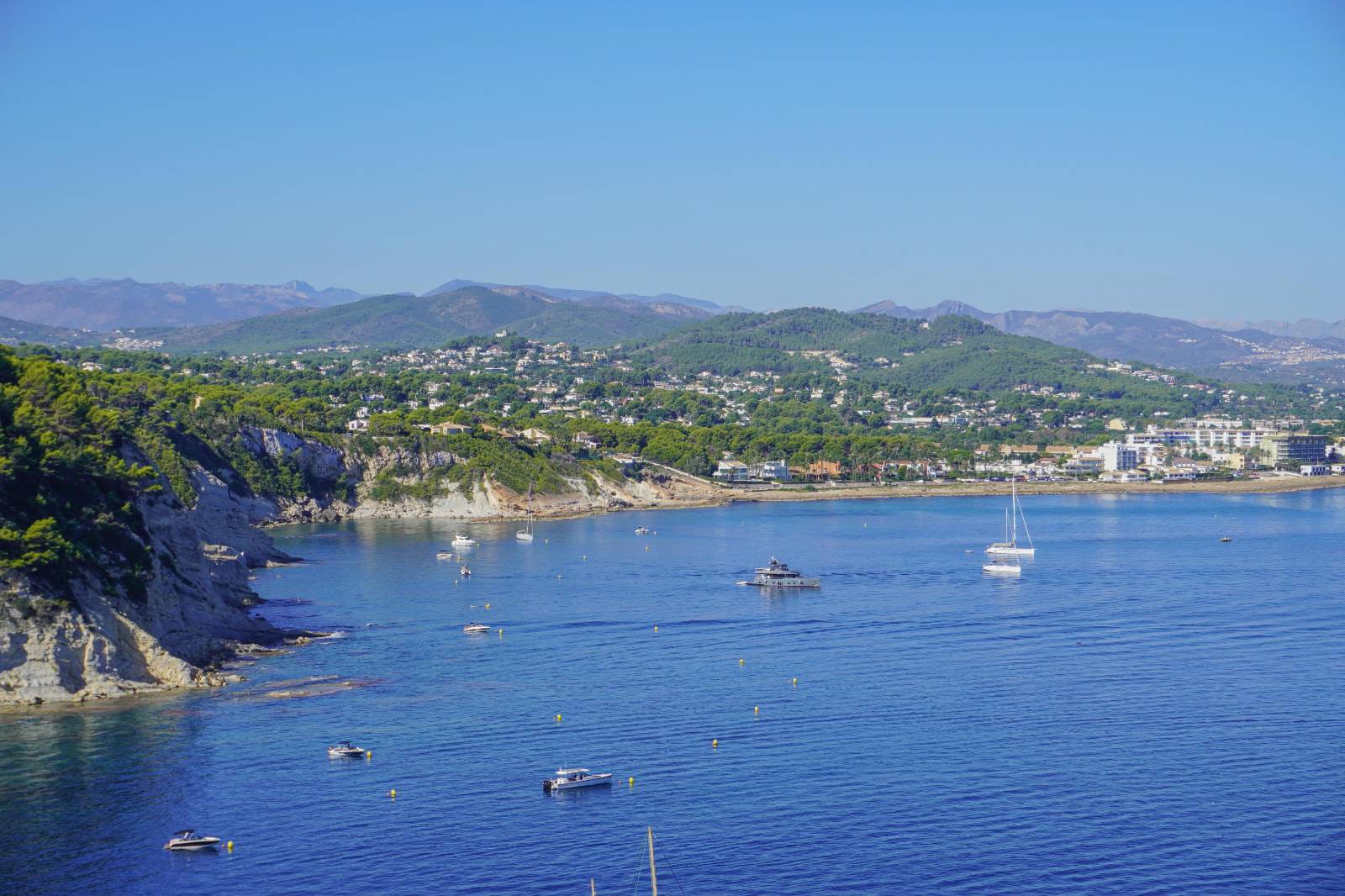 Vista panorámica de la bahía de Cala Blanca en Jávea, mostrando las aguas cristalinas del Mediterráneo con embarcaciones fondeadas y el relieve costero de pinos y acantilados.