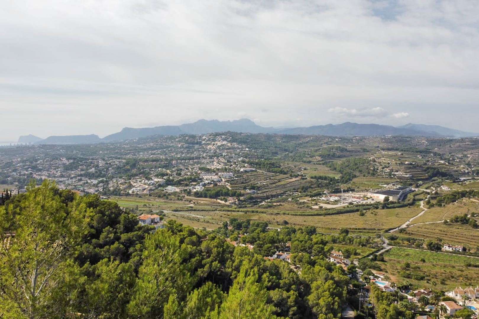 Vista panorámica elevada desde la parcela hacia el valle de Moraira, destacando la densa vegetación de pinos y las siluetas montañosas al fondo.