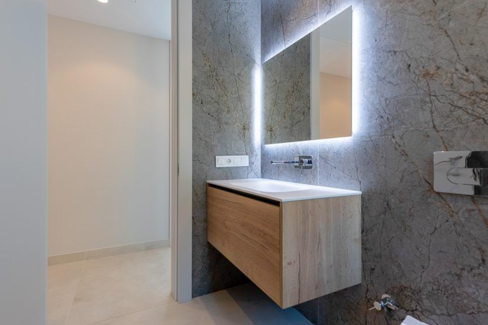 Aseo de cortesía con mueble suspendido en madera, lavabo integrado y espejo retroiluminado sobre pared de gres porcelánico con efecto mármol gris.