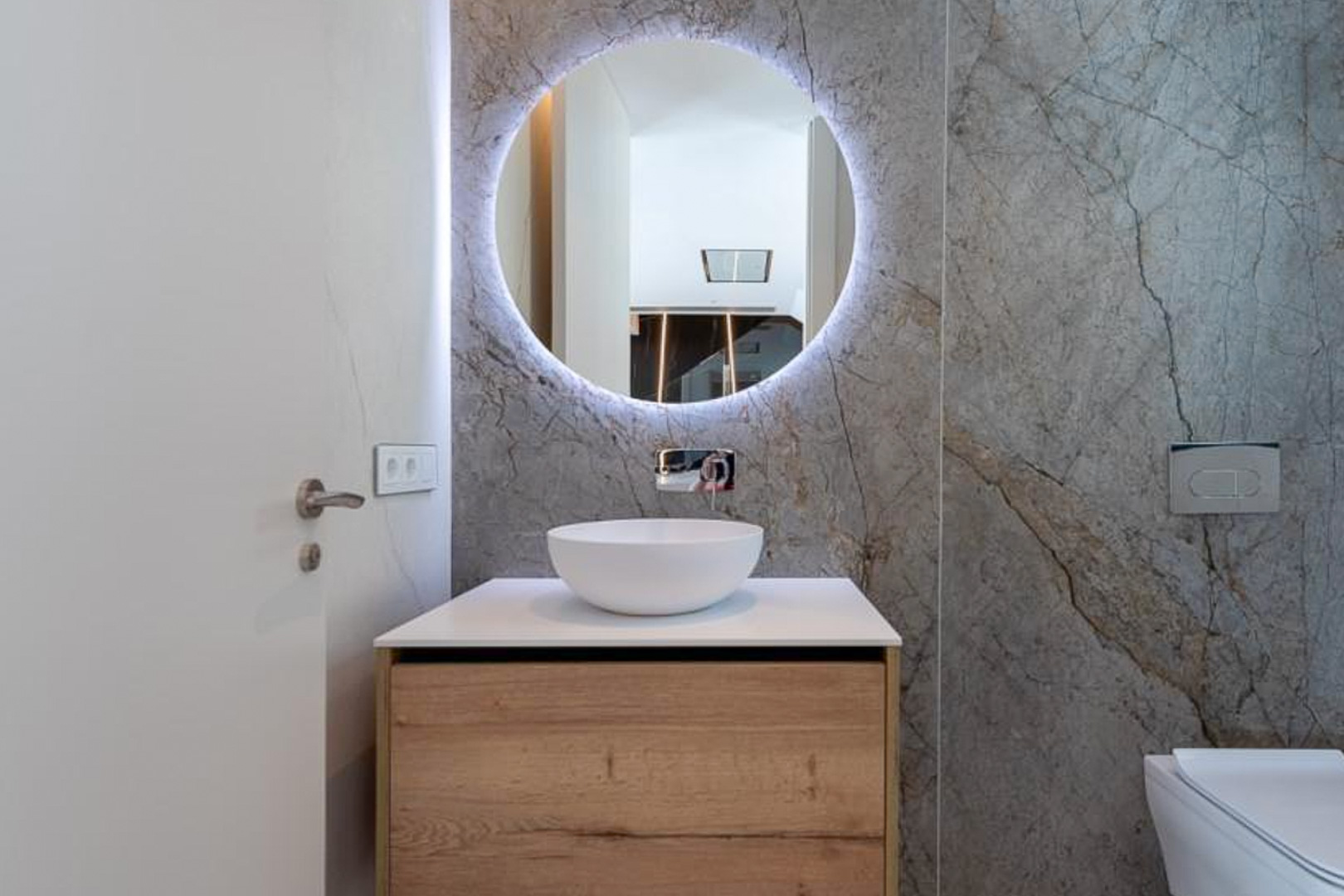 Aseo de cortesía con revestimiento de mármol gris, espejo circular retroiluminado y mueble de madera con lavabo sobre encimera y grifería empotrada.