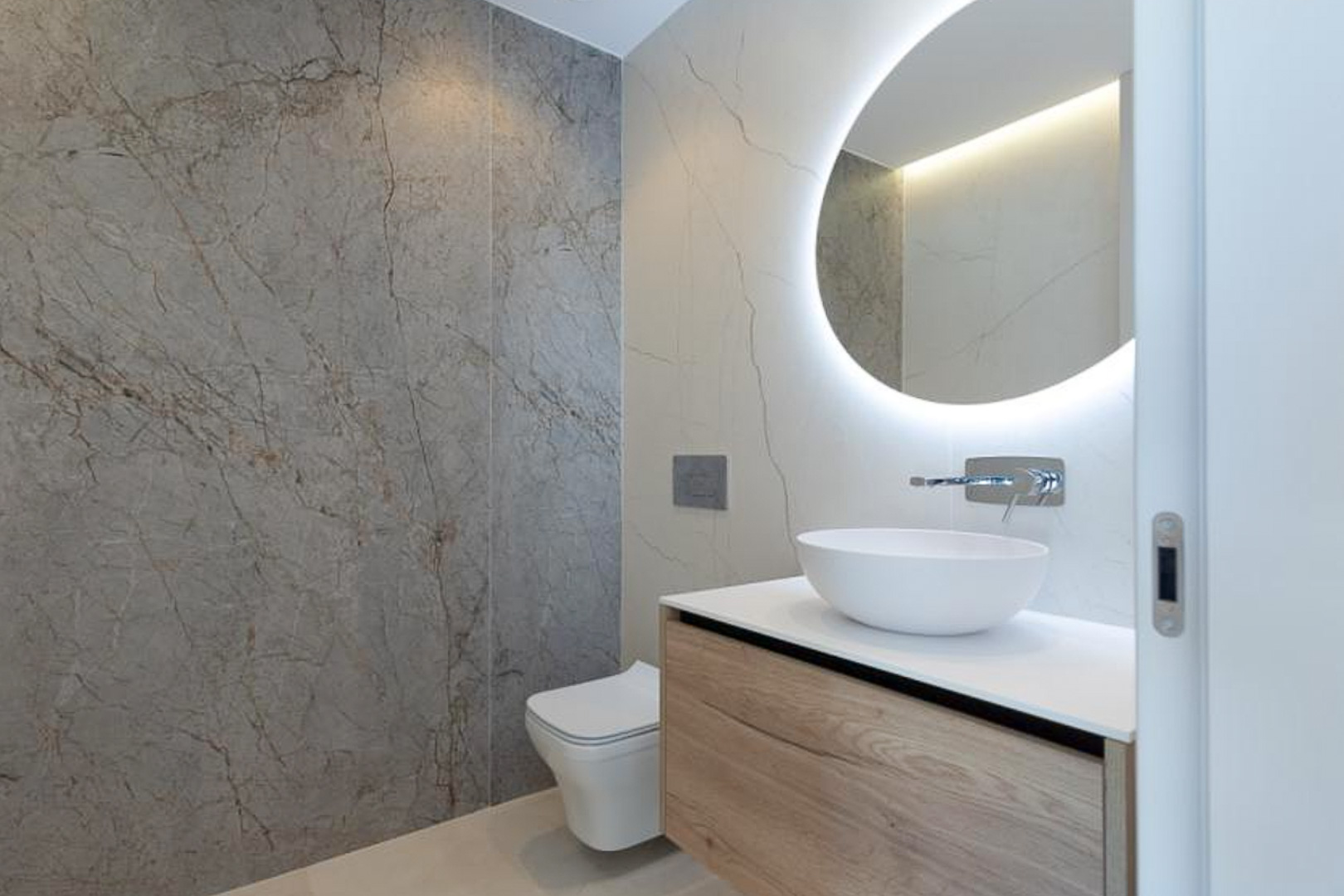 Aseo de diseño con lavabo sobre encimera, mueble suspendido de madera y espejo circular retroiluminado sobre paredes de gres porcelánico efecto mármol.