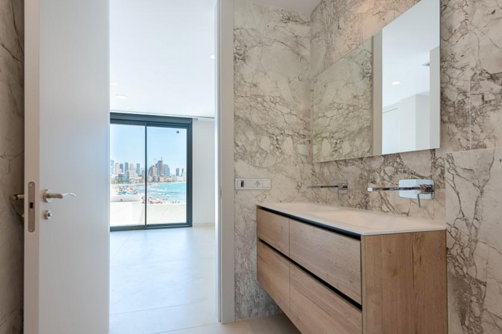 Baño de diseño con revestimiento de mármol, mueble de madera de doble seno y grifería empotrada, con vistas al mar a través del dormitorio.